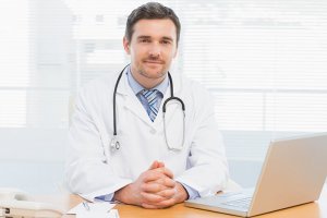 Portrait of a smiling male doctor with laptop sitting at desk in medical office